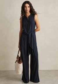 Marineblauwe mouwloze jumpsuit met een knoopsluiting aan de voorkant, een strikdetail in de taille en een wijde pijpdesign; gecombineerd met een bruine en crèmekleurige schoudertas.