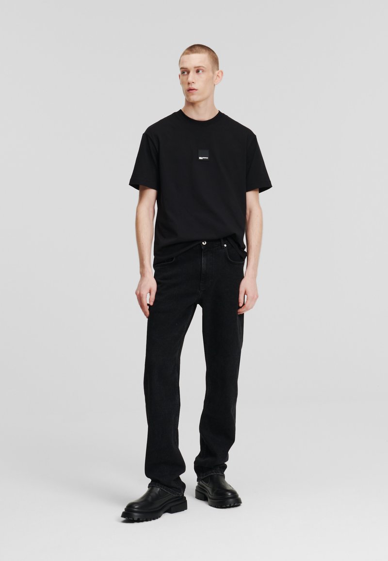 Camiseta negra con un logo cuadrado en el lado izquierdo del pecho, combinada con unos vaqueros negros de pierna recta y botas negras de suela gruesa.