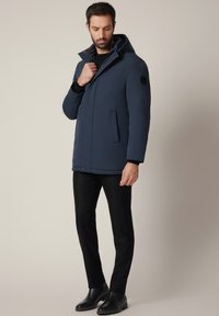 Mężczyzna poprawiający kołnierz granatowej zimowej kurtki z kapturem, ubrany w czarne spodnie i czarne skórzane buty, stojący na jednolitym beżowym tle.