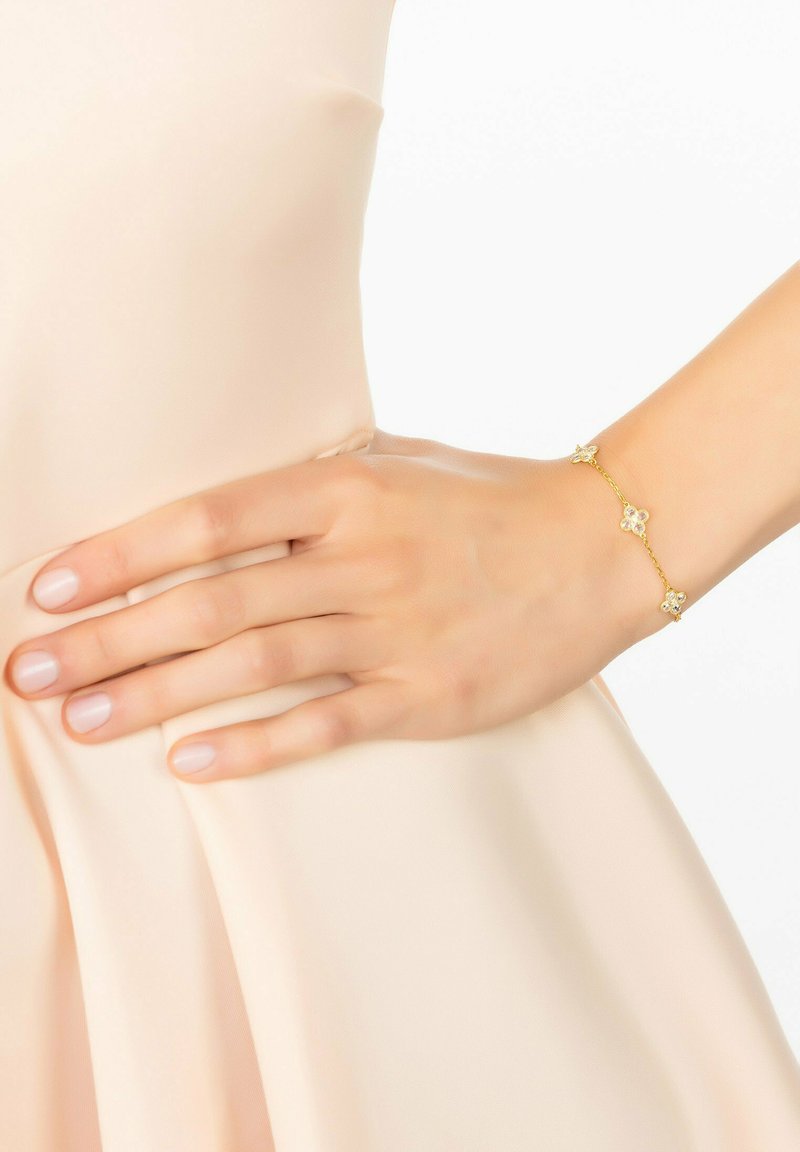 Arany karkötő virágos díszítéssel, három dekoratív virággal. Finom láncfrastruktúra, amely egy puha, világos szöveten nyugvó kézen viselhető.