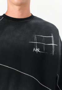 Schwarzer Pullover mit Rundhalsausschnitt, der weiße gestickte Linien und ein geometrisches Design auf der Brust aufweist. Kontrastierende Nähte.