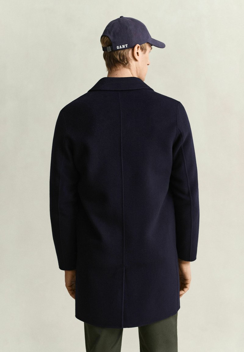 Homme portant un manteau bleu marine et une casquette bleu marine avec le texte "GANT", vu de dos sur un fond clair uni.