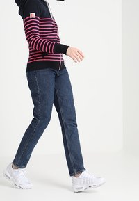 Felpa a strisce navy e rosa con zip frontale, abbinata a jeans blu e scarpe da ginnastica bianche, mostrata su uno sfondo neutro.