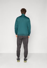 Teal sportsjakke med ståkrage, kombinert med svarte smale bukser. Glatt tekstur, elastiske mansjetter og elastisk midje, på en nøytral bakgrunn.
