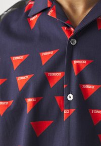 Tamsiai mėlynos spalvos trumpomis rankovėmis mygtukinė marškinėliai su raudonais trikampiais raštais, kuriuose yra žodis "Fiorucci" baltai. Paprastas apykaklė, vienas mygtukas.