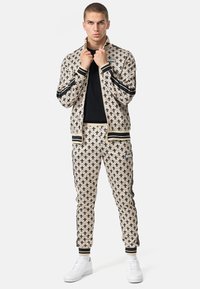 Tan tracksuit with black floral patterns, featuring a zip jacket and elastic waist pants. Accented with black stripes and white sneakers.