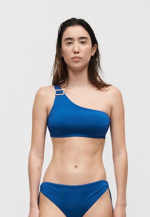 Γυναίκα με βρεγμένα μαλλιά που φοράει μπλε μπικίνι με έναν ώμο και πόρπη, καθώς και ταίριαστο κάτω μέρος, σε μονόχρωμο φόντο.