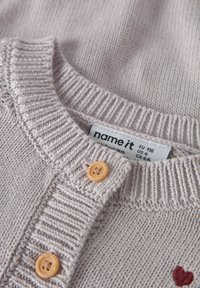 Hellgraue Strickjacke mit geripptem Kragen, zwei Holzknöpfen und einem kleinen roten Herzdetail. Etikett mit Größenangabe sichtbar.