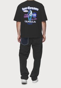 Camiseta negra con estampado gráfico que presenta un cucurucho de helado y texto, combinada con jeans negros y zapatillas blancas. Los detalles notables incluyen un bolsillo de mezclilla.