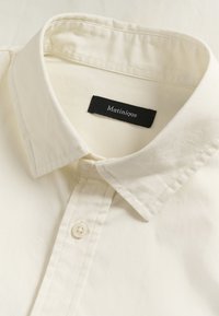 Chemise en coton crème avec un col plié, comportant une étiquette noire portant l'inscription « Matinique » et un bouton blanc au niveau du col. Texture lisse.