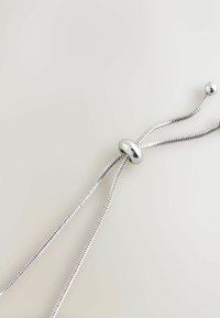 Srebrny metaliczny naszyjnik z elastycznym, żebrowanym łańcuchem, z gładką owalną kulką jako zapięciem, na neutralnym tle.