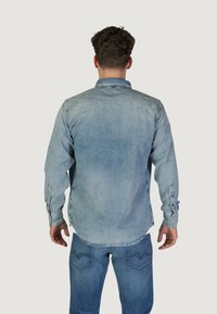 Camicia di jeans azzurro chiaro con maniche lunghe, chiusura a bottoni e colletto classico. Texture invecchiata e dettagli di cucitura sono ben visibili.