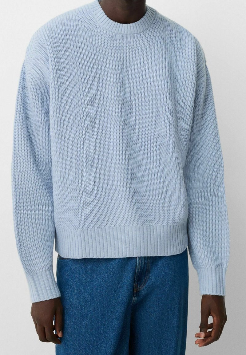 Mann in einem hellblauen gerippten Strickpullover und blauen Jeans, der vor einem schlichten Hintergrund steht.