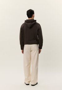 Persona in piedi che indossa una felpa con cappuccio marrone scuro e pantaloni beige larghi contro uno sfondo chiaro e uniforme.