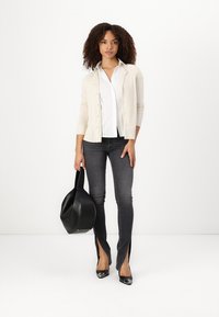 Beige Cardigan über weißem Hemd, kombiniert mit dunklen Jeans mit seitlichen Schlitzen. Das Modell hält eine schwarze Tasche und trägt schwarze Absätze. Insgesamt glatte Texturen.