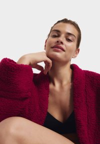 Bata de felpa texturizada burdeos con cuello de chal. La modelo posa con la barbilla apoyada en la mano, vistiendo un top de bikini negro.