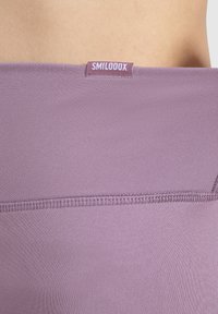 Leggings deportivos de color púrpura claro con una textura suave y elástica, y una etiqueta de marca visible etiquetada como "SMILOOOX" en la cintura.