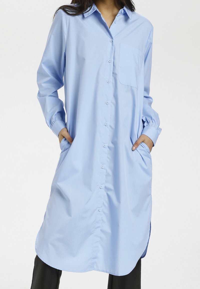 Femme portant une robe chemise longue bleu clair avec manches longues et poches latérales, debout les mains dans les poches.