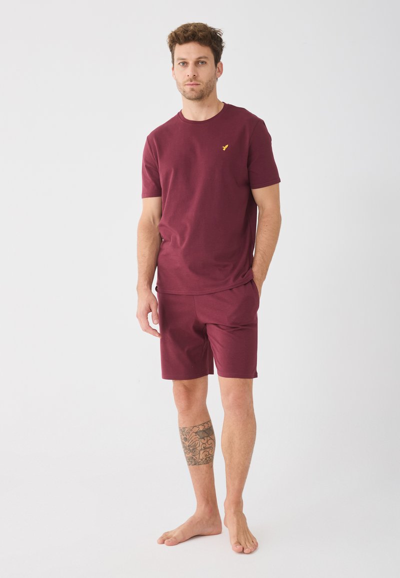 Mørkerød t-shirt med korte ærmer og et gult logo, parret med matchende mørkerøde shorts. Bomuldsstof, afslappet pasform, minimalistisk design og blød tekstur.