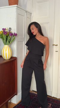 Γυναίκα με μαύρο τοπ με έναν ώμο και φαρδιά παντελόνια στέκεται δίπλα σε λευκή πόρτα, κοντά σε ξύλινη ντουλάπα με βάζο λουλουδιών με κίτρινες ρίγες.