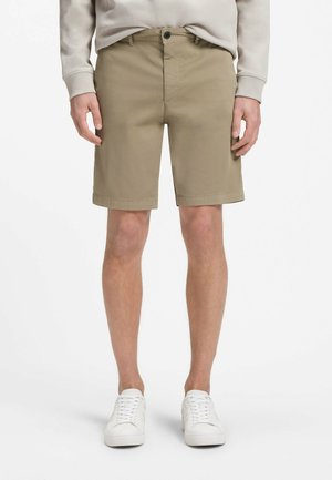 Mann trägt beige kniehohe Shorts, einen hellgrauen Pullover und weiße Turnschuhe, steht vor einem schlichten weißen Hintergrund.