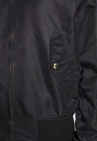 Veste noire avec fermeture éclair, poche à rabat inclinée avec bouton-pression doré et ceinture en maille côtelée sur le côté droit.