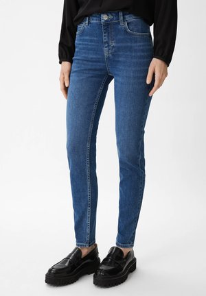 Schmale blaue Jeans mit hoher Taille, frontalen Taschen und dezenten Used-Effekten. Kombiniert mit schwarzen glänzenden Loafern für den Kontrast.