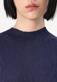 Top tricotat texturat de culoare albastru marin, cu un guler înalt, ribat. Țesătura prezintă modele subtile și inegale, care adaugă adâncime designului.