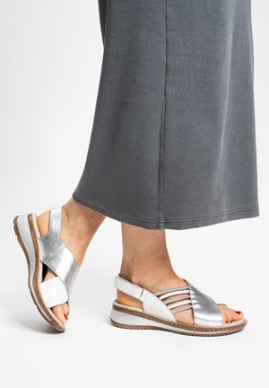 Feet wearing silver metallic open-toe sandals with straps and white soles, paired with a gray ankle-length skirt.
