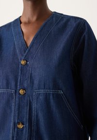 Ciemnoniebieska jeansowa kurtka z dekoltem w kształcie litery V, zapinana na cztery guziki, z dwoma przednimi kieszeniami. Charakteryzuje się kontrastowymi przeszyciami i luźnym krojem.