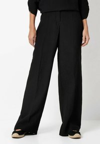 Black wide-leg trousers made of textured fabric, featuring a high waist and straight cut. Worn with espadrille-style shoes.