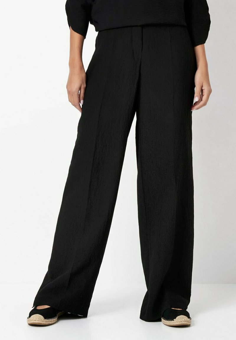 Black wide-leg trousers made of textured fabric, featuring a high waist and straight cut. Worn with espadrille-style shoes.