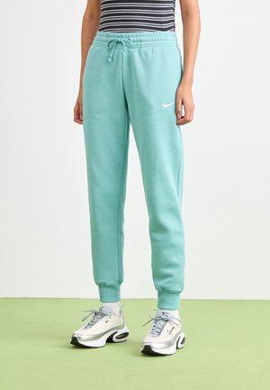 Persoon die een lichtblauwe Nike joggingbroek, witte Nike sneakers, een zwart-wit gestreept shirt draagt, staand op een groene vloer tegen een witte achtergrond.