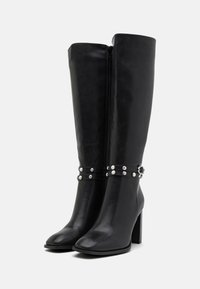 Botas negras de cuero suave hasta la rodilla, con cremallera lateral y adornadas con tachuelas plateadas en dos correas alrededor del tobillo.