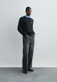 Μαύρος πουλόβερ με μικρό πορτοκαλί έμβλημα, ανοιχτό μπλε πουκάμισο με γιακά από κάτω, γκρι φαρδιά παντελόνια και μαύρα παπούτσια σε ουδέτερο φόντο.