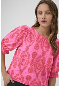 Blusa rosa ricamata con maniche a sbuffo, motivi ovali e motivi floreali. Tessuto morbido con dettagli testurizzati.
