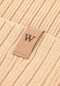 Beige geribbelde gebreide stof met een klein tan leren label waarop een zwart "W" logo staat. De textuur is zacht en rekbaar, met verticale lijnen.