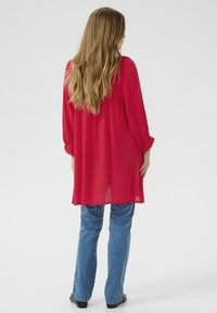 Blouse rouge à manches longues en tissu léger, associée à un jean bleu. La blouse est légèrement oversize, avec une coupe ample et un ourlet droit.