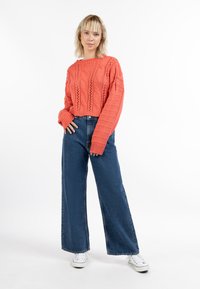 Korallfarbener, kabelstrickfarbener, verkürzter Pullover mit weiten Ärmeln, kombiniert mit hoch taillierten blauen, weiten Jeans und weißen Sneakers. Schlichter weißer Hintergrund.