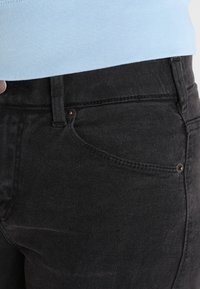 Schwarze Jeans aus Denim mit glatter Textur, ausgestattet mit einer Fronttasche und einem braunen Knopfakzent. Kombiniert mit einem hellblauen gerippten Oberteil.
