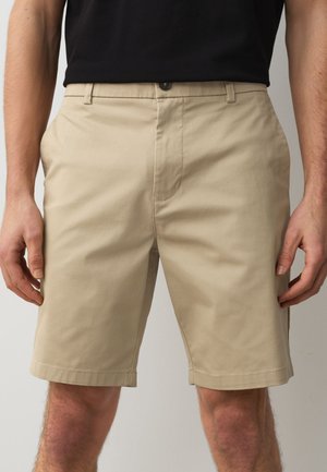 Mand iført beige knælange shorts med frontknap og lynlås, kombineret med en sort skjorte, stående med hænderne afslappet ned langs siderne.