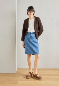 Cardigan beige et duveteux, t-shirt blanc, jupe crayon en denim et sandales à plateforme brunes. Salle avec un sol en bois et des murs neutres.