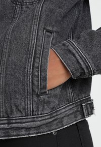 Šedá džínová bunda s bílým prošíváním, se boční kapsou, detaily na manžetách a mírně opraným spodním lemem.