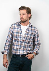 Koszula w kratę w jasnoniebieskich, białych i pomarańczowych odcieniach, zestawiona z białym t-shirtem i ciemnymi dżinsami. Srebrny zegarek na nadgarstku.