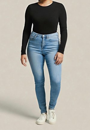 Frau trägt hellblaue, hoch taillierte Skinny-Jeans, schwarzes Langarmoberteil und weiße Turnschuhe, steht vor einem schlichten Hintergrund.