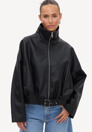 Femme portant une veste en cuir noire zippée avec un col montant et une ceinture, associée à un jean bleu clair, debout devant un fond uni.