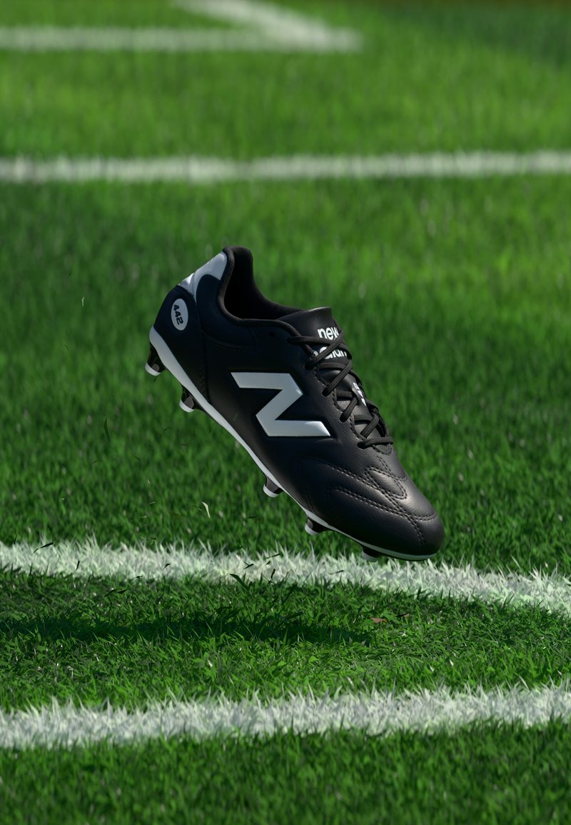 Chaussure de football noire New Balance avec logo blanc, flottant au-dessus d'un gazon artificiel vert marqué de lignes blanches.