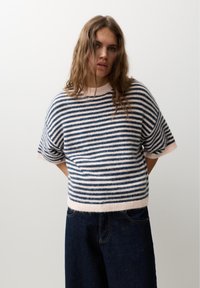 Kvinde med langt brunt hår iført en kortærmet marineblå og hvidstribet sweater og mørkeblå jeans, stående foran en ensfarvet baggrund.