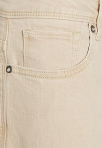 Světlé béžové džínové kalhoty se zadní kapsou s kontrastním prošitím a kovovým knoflíkem na pásku.