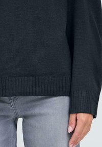 Mörkgrå stickad tröja med ribbad nederkant och mudd. Mjuk textur med avslappnad passform, ihop med ljusgrå jeans.